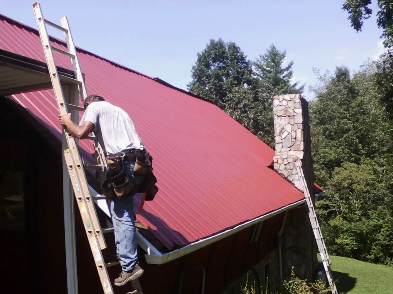 Roofer on a ladder beside a red metal roof and stone chimney in Spicer